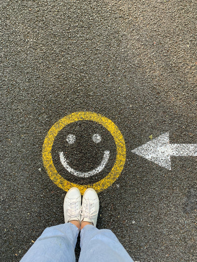 Someone wearing blue jeans and white trainers stood on concrete next to a smiley face