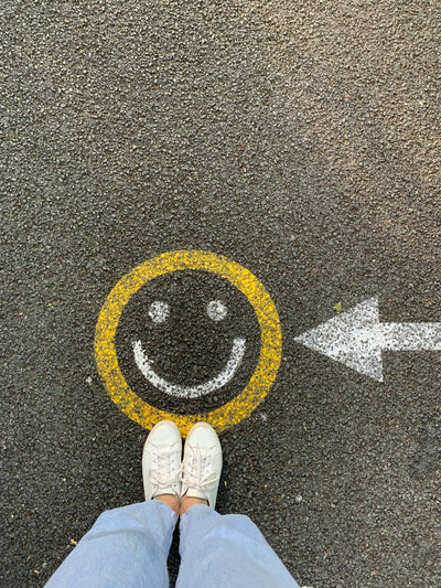 Someone wearing blue jeans and white trainers stood on concrete next to a smiley face