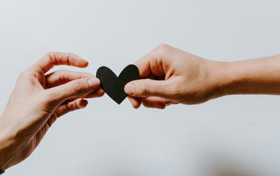 two hands meeting in the middle joined by a black paper heart each holding one end