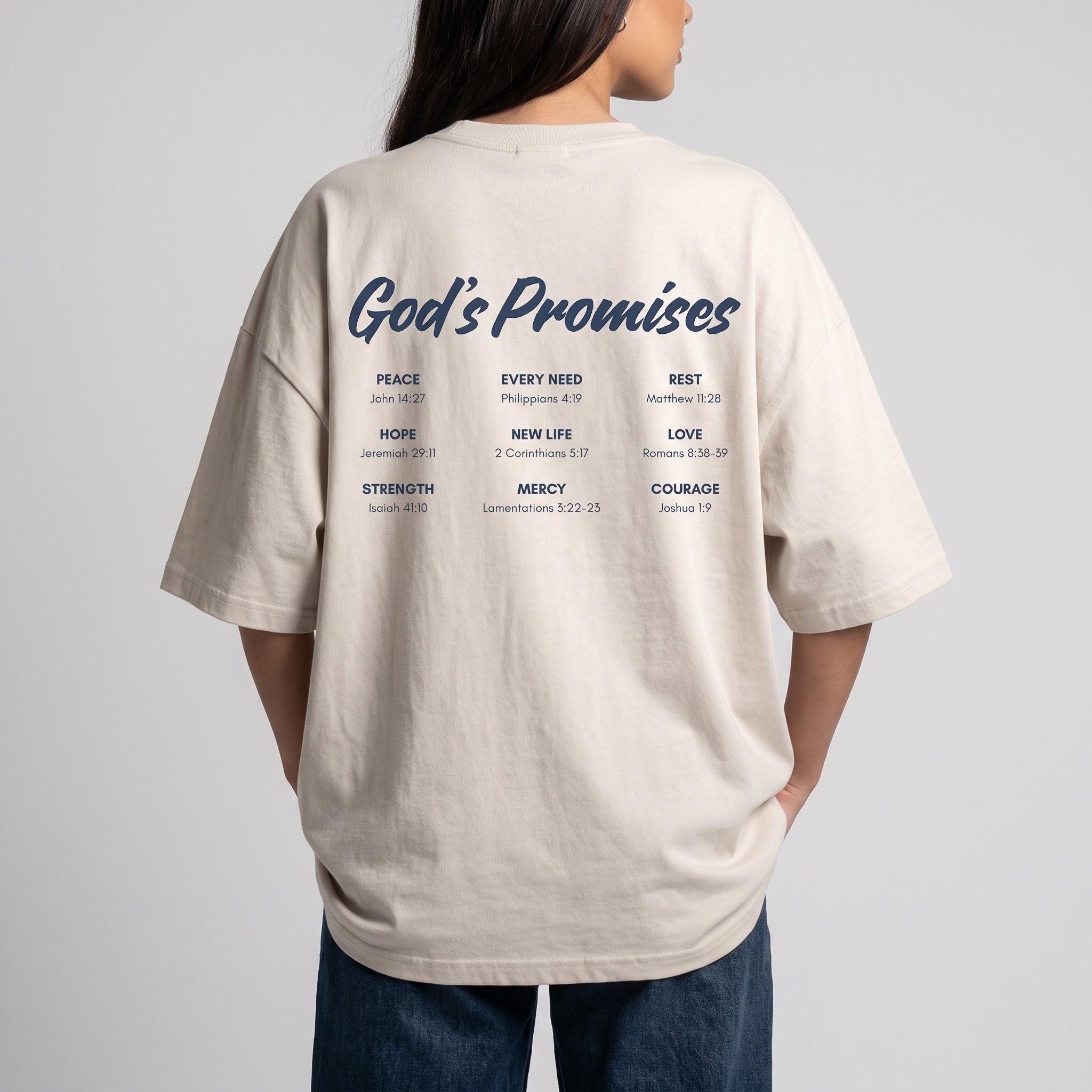 A woman shown from the back wearing an oversized off-white t-shirt with a dark blue graphic print on the back that reads 'God's Promises' followed by various biblical promises and verses. She is also wearing blue jeans, set against a plain light gray studio background.