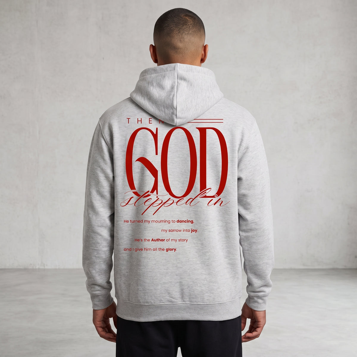 A man viewed from behind wearing a light gray hoodie with a large red graphic text print on the back reading 'GOD stepped in', set against a neutral gray textured wall.