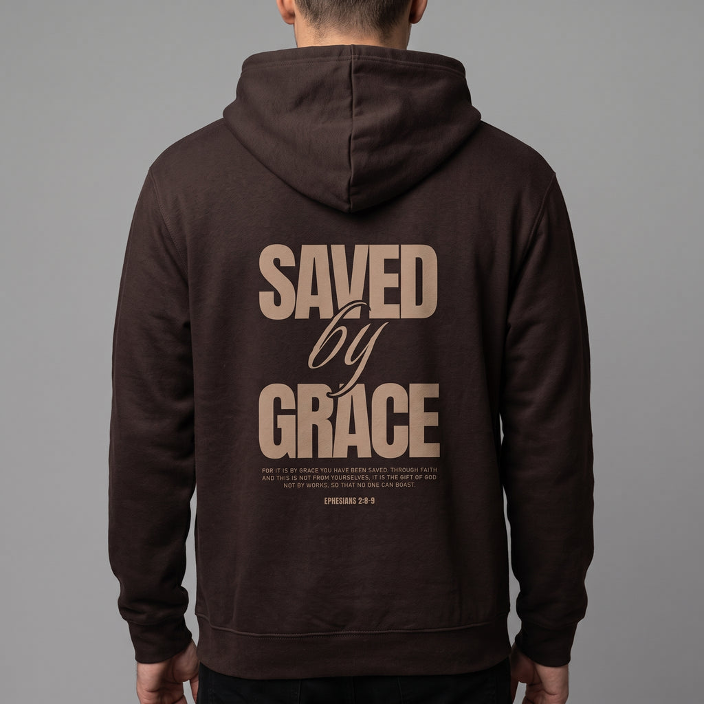 A man viewed from behind wearing a dark brown hoodie with a large tan graphic print on the back reading 'SAVED by GRACE' and smaller scripture text below, against a plain gray studio background.