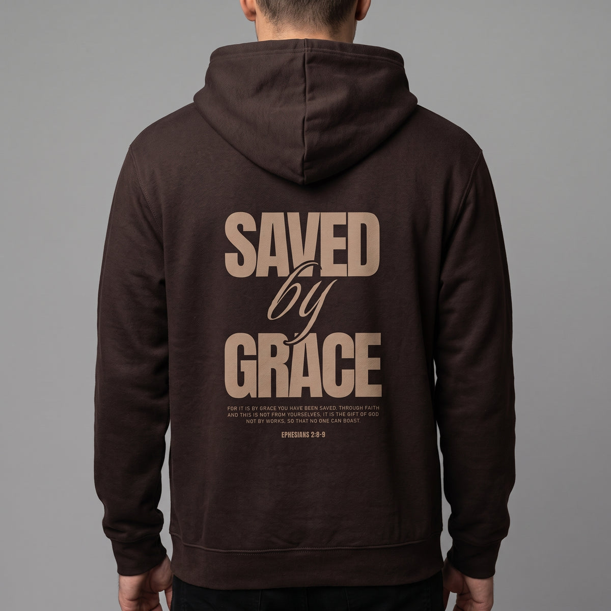 A man viewed from behind wearing a dark brown hoodie with a large tan graphic print on the back reading 'SAVED by GRACE' and smaller scripture text below, against a plain gray studio background.