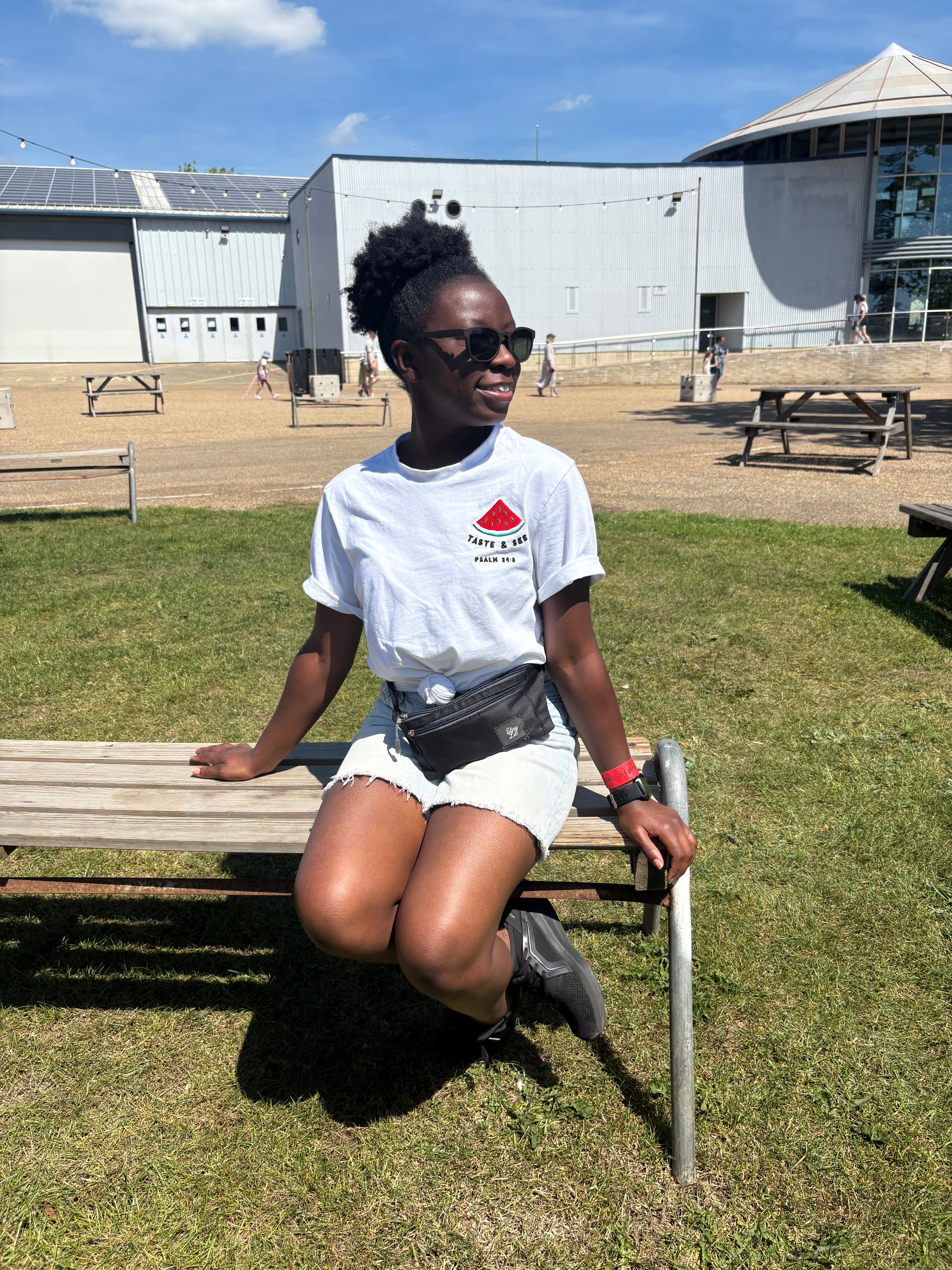 Person sitting on a bench outdoors with a building and blue sky in the background in Taste and see t-shirt by Stay Lit Apparel