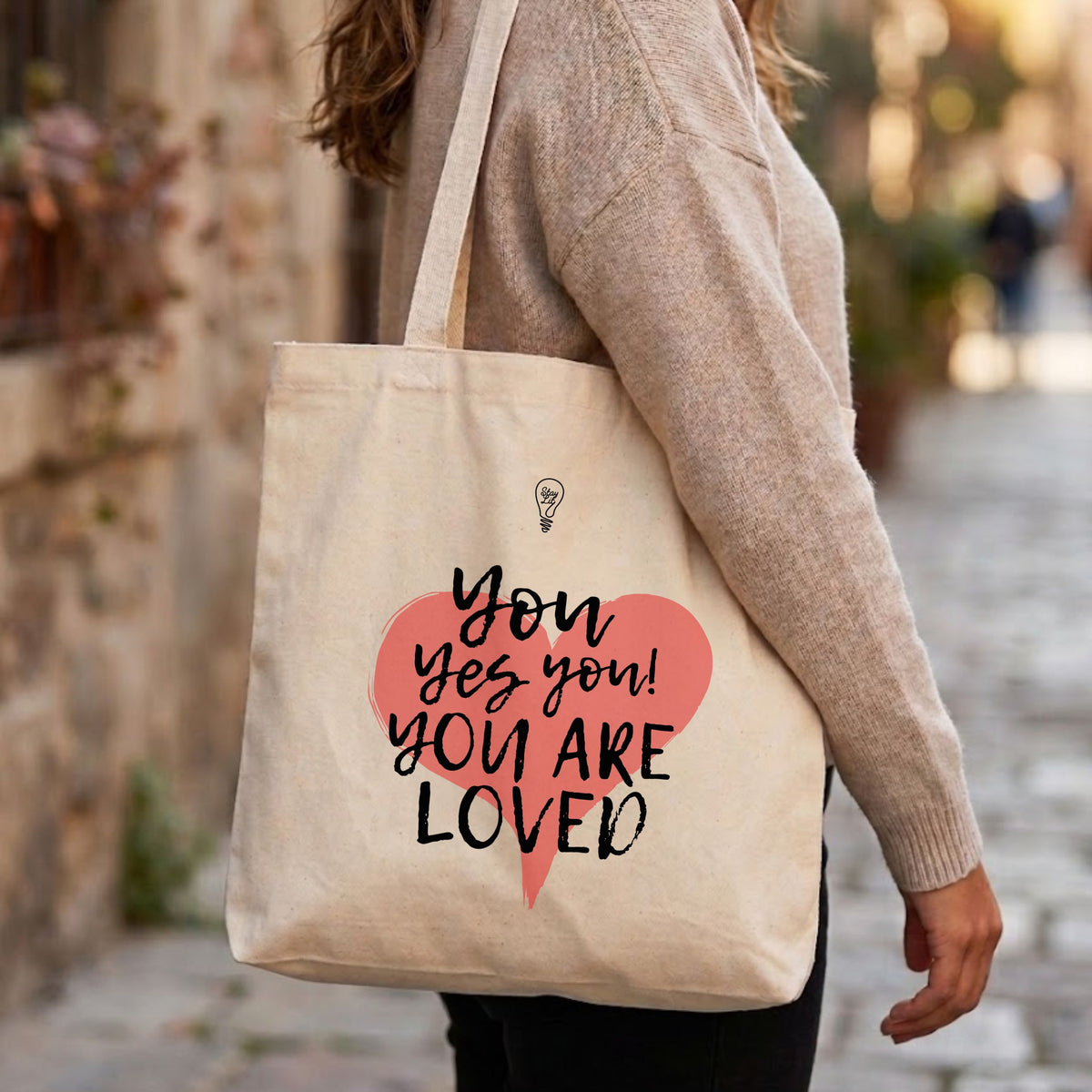 A person wearing a beige knit sweater carries a cream canvas tote bag with a pink heart and black text reading 'You yes you! YOU ARE LOVED' on a blurred outdoor street background.